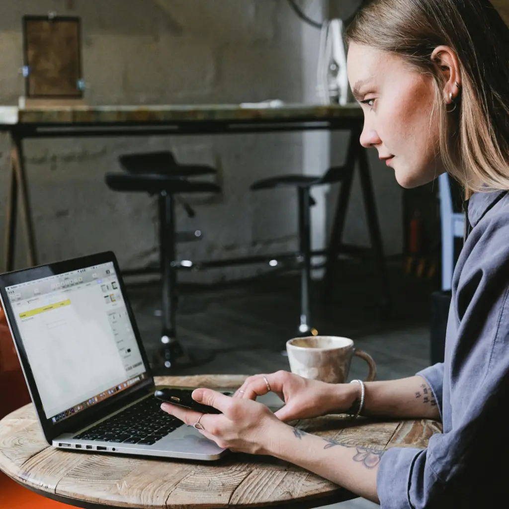 Une personne est assise à une table ronde en bois dans une pièce confortable, travaillant sur un ordinateur portable tout en tenant un smartphone.