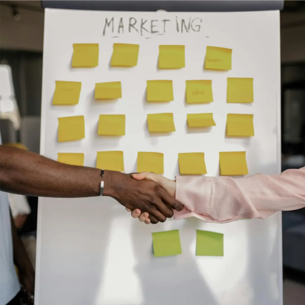 Deux personnes se serrent la main devant un tableau blanc avec plusieurs notes autocollantes jaunes sous le titre "MARKETING".