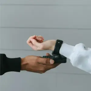 La photo montre deux mains de personnes différentes en interaction. La main de gauche est celle d'une personne à la peau foncée, portant un pull noir, et elle tient un terminal de paiement. La main de droite est celle d'une personne à la peau claire, portant un sweat-shirt gris clair et une montre noire. Cette main est en train d'effectuer un paiement sans contact en approchant son poignet, où se trouve la montre, du terminal de paiement. L'arrière-plan est flou avec des teintes gris clair.